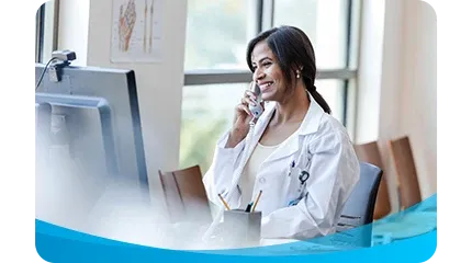 A smiling female doctor or medical professional wearing a white lab coat is sitting at a desk and talking on a landline phone next to a computer monitor.